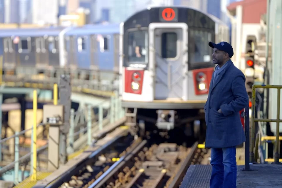 Subway train pulling into elevated outdoor platform in New York City with one passenger waiting.